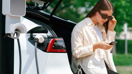 Young woman use smartphone to pay for electricity at public EV car charging station green city...