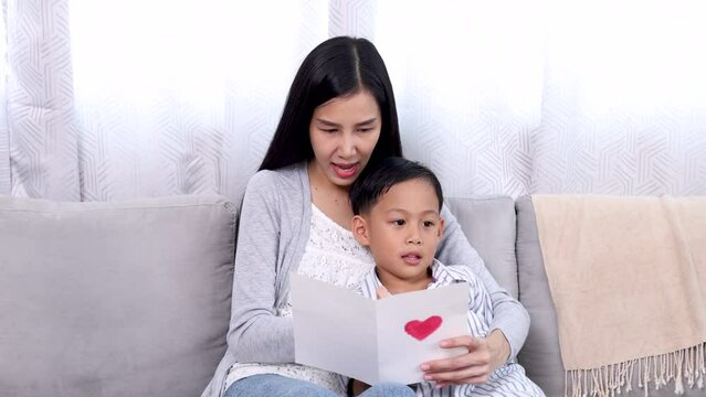 Young Asian single mom entrepreneur surprise lovely card from adorable cute son in living room, mother working at home and take care boy, mother's day concept kids and female love bonding relationship