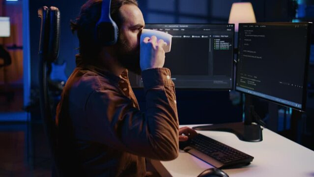 Developer listening music and drinking coffee while doing programming during remote job shift in home office. IT expert enjoying audio podcast and hot beverage, working on PC, camera B