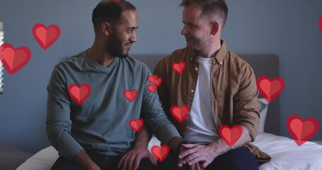 Image of red hearts over diverse male couple sitting on bed