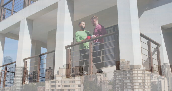 Image of cityscape over diverse male couple standing on balcony and drinking coffee - Powered by Adobe