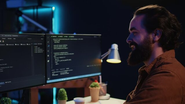 Smiling freelancing developer in home office doing coding on computer at desk. Cheerful IT expert doing contracted job from apartment, writing lines of code at workstation, camera B close up