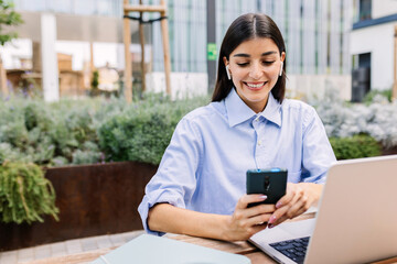 Young business woman writing message on mobile phone while working on laptop sitting outside of office building.