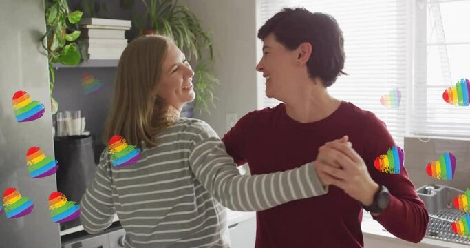 Animation of pride rainbow hearts over happy caucasian lesbian couple dancing in kitchen