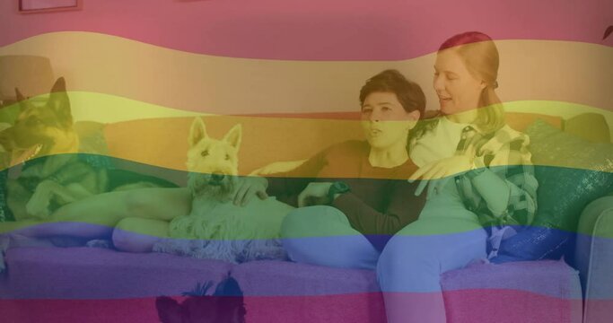 Animation of pride rainbow flag over happy caucasian lesbian couple relaxing on couch with pet dogs