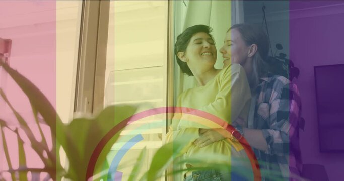 Animation of pride rainbow flag and rings over happy caucasian lesbian couple embracing at home