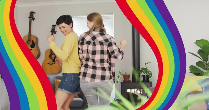 Animation of pride rainbow bands over happy caucasian lesbian couple dancing at home