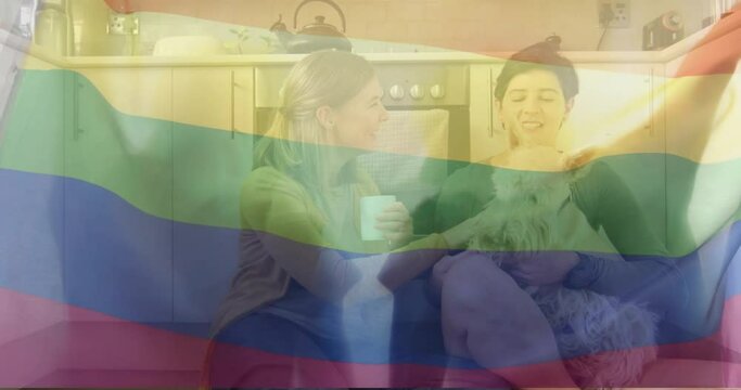 Animation of pride rainbow flag over happy caucasian lesbian couple sitting with pet dog in kitchen