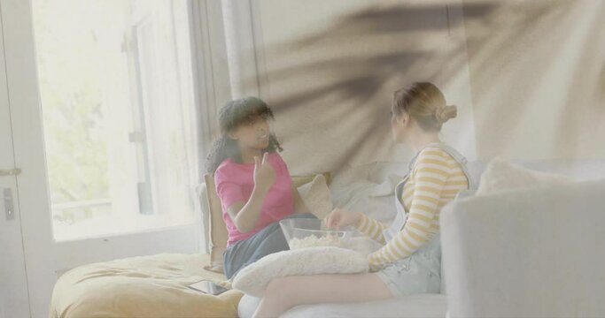 Animation of plant leaves over two happy diverse teenage girls talking and eating popcorn at home