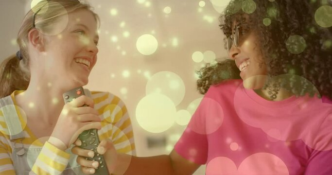 Animation of light spots over two happy diverse teenage girls miming singing with tv remote control