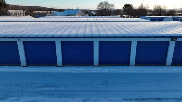 Aerial trucking shot of Rental Garage Units with blue doors at snowy winter day. Snow covered roof and street at sunny day. American suburb area, USA.