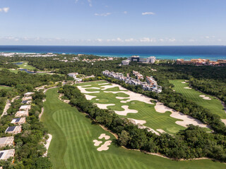 Drone view of golf course and green tropical vegetation on a sunny day with hotel resorts and blue Caribbean Sea in the background in Tulum