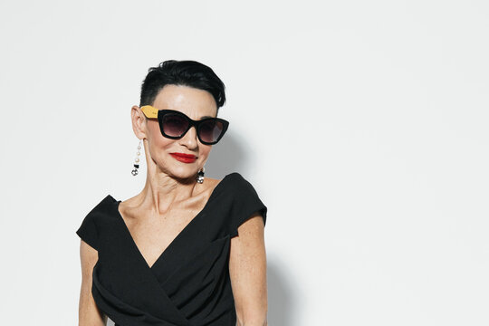 Confident woman in elegant black dress and sunglasses posing with hands on hips in front of white wall