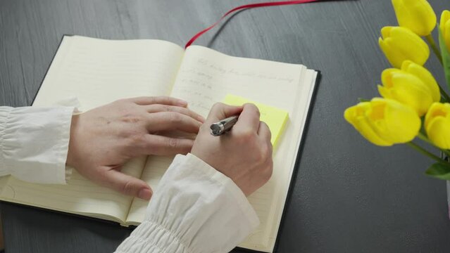 Hands of a woman writing notes in her agenda and on post it, at a table decorated with yellow tulips. Lifestyle concept. Close-up.