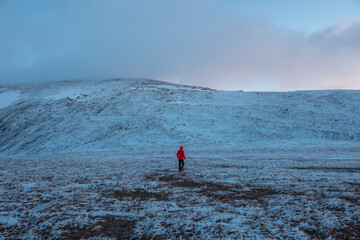 Man in red jacket in hood on snow-covered stony pass against snowy mountain wall with top in low...