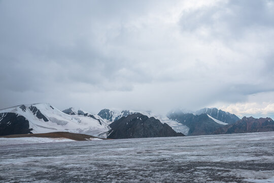 Dramatic vast misty top view from ice pass to big glacier tongue among sharp rocks and large snow-capped mountains in rainy low clouds. Dark atmospheric mountain silhouettes in rain in gray cloudy sky