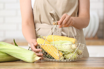 Woman holding basket with fresh corn cobs on wooden table in kitchen