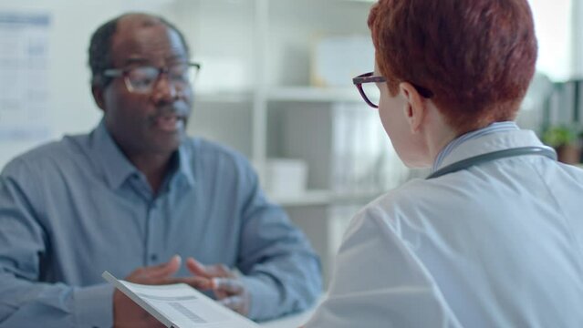 Female doctor in white coat reading medical records and asking male patient about complaints during appointment in clinic. Focus on foreground