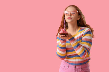 Beautiful young woman blowing out candle on birthday cupcake against pink background