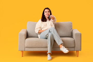 Obraz premium Beautiful young happy woman sitting on sofa and watching TV against yellow background