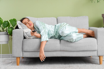 Beautiful young bored woman lying on sofa and watching TV near green wall