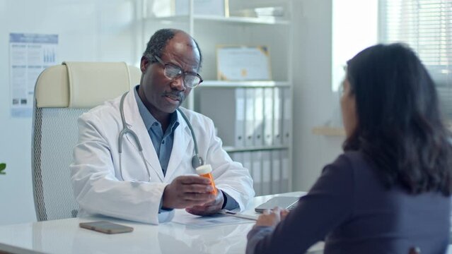 Mature African American doctor giving pills to female patient and explaining the dose and treatment plan during medical consultation in clinic