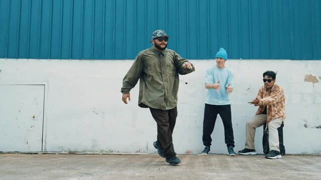 Skilled hipster doing cart wheel while multicultural friends encourage him. Hip-hop dancer moving foot step to music surrounded by group of hipster at wall near street. Outdoor sport 2024. hiphop.