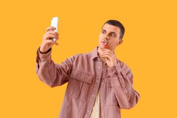 Young man with electronic cigarette taking selfie on yellow background