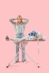 Panicked mature woman with pile of laundry on ironing board against pink background