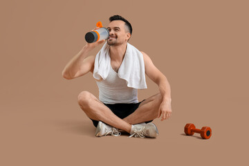 Handsome young man in sportswear with bottle of water and dumbbell sitting on brown background