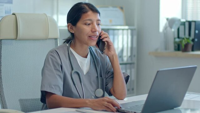 Latin female physician wearing scrubs sitting at desk with laptop and having talk with patient on mobile phone during workday