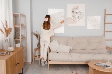 Young woman hanging stylish painting on light wall in living room, back view