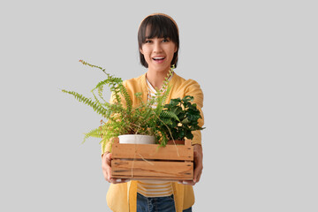 Beautiful young happy female gardener holding wooden box with houseplants on grey background