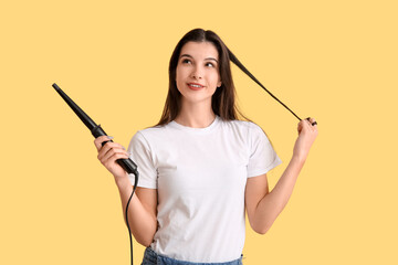 Beautiful young happy woman with curling iron on yellow background