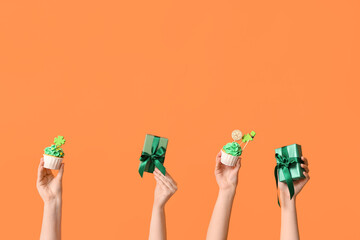 Female hands with tasty cupcakes and gift boxes for St. Patrick's Day on orange background