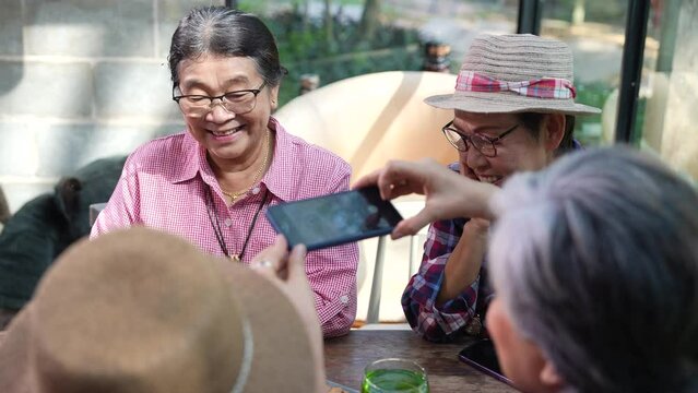 Group of Asian seniors, pensioners, elderly, and retirement friends laughing talking sharing idea spend free time get together at cozy restaurant, happy using smartphone friendly technology learning