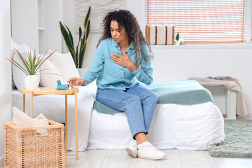 Young African-American woman taking asthma inhaler from table in bedroom