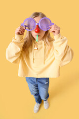 Little girl in funny disguise with party blower on yellow background. April Fools' Day celebration