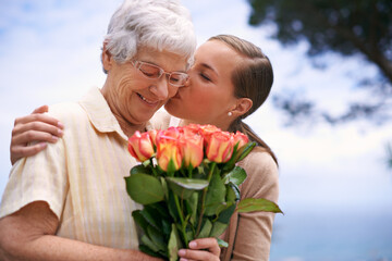 Senior woman, daughter and kiss with flowers, hug and care for love, bonding and reunion at family home. People, elderly mom and mothers day celebration with connection, gift and bouquet in garden - obrazy, fototapety, plakaty