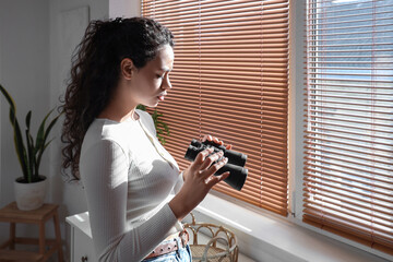 Young African-American woman with binoculars near window at home