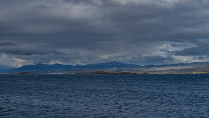 The coastline of the Tierra del Fuego archipelago is visible from the Beagle Channel. The Andes mountains against a cloudy sky. Ripples on the blue water of the strait. Argentina.