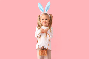 Cute little girl in bunny ears with basket of chocolate Easter eggs on pink background
