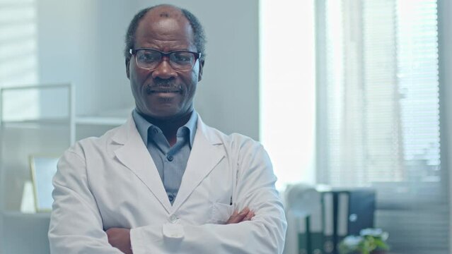 Senior African American general practitioner in white coat holding arms crossed and smiling at camera when working in clinic. Video portrait