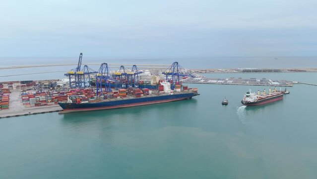 Expansive view of an industrial seaport showing cargo terminals, container ships at berth, multiple cranes against a backdrop of calm seas and a slightly overcast sky. Activity in hub of commerce 