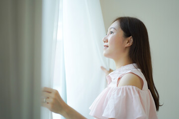 Happy Beautiful young Asian woman opening window curtain in the morning. Portrait of Pretty Asian...