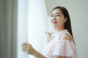 Happy Beautiful young Asian woman opening window curtain in the morning. Portrait of Pretty Asian young woman looking the sun rise outside through the window peacefully, Asian girl portrait close up.