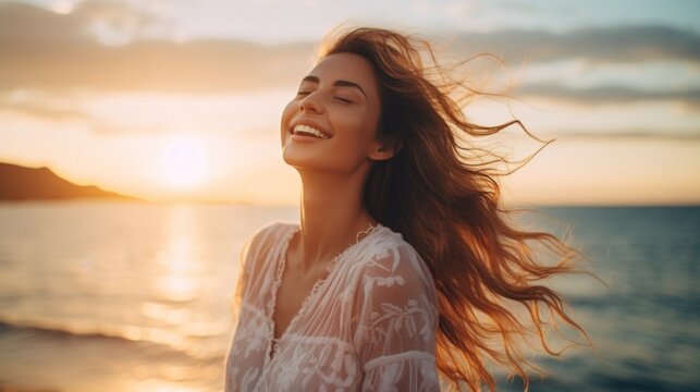 A happy beautiful young smiling woman with hair flying in the wind, having fun on the beach with her eyes closed at sunset. Golden Hour, Pleasure, Joy, Travel, Summer, Vacation concepts.