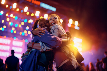 Obraz na płótnie Canvas Cheerful female friends embracing in front of music stage during summer festival at night.