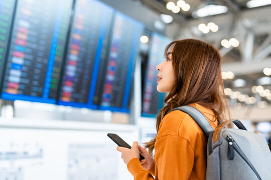 Young asian woman in international airport, using mobile smartphone and checking flight at the flight information board