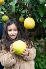 Young girl with a beaming smile holding a large pomelo, surrounded by green foliage.